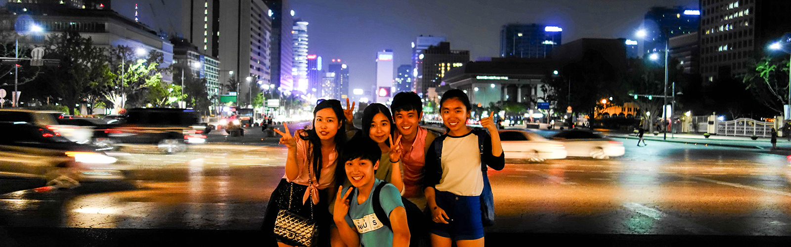 Five students posed near a busy road in Seoul at night, with brightly-lit buildings in the background.